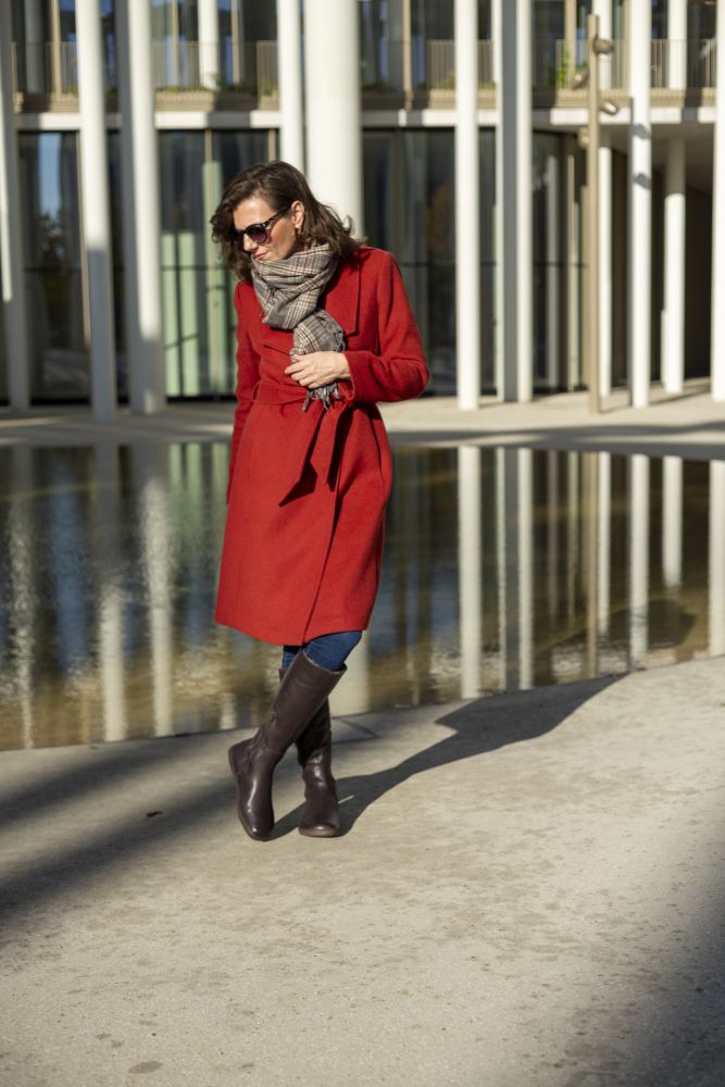 Eine Frau steht an einem sonnigen Tag neben einer reflektierenden Oberfläche vor einem modernen Gebäude mit hohen Säulen. Sie trägt eine Sonnenbrille und einen grauen Schal. Ihr stilvolles Ensemble besteht aus den QATCH Winter Brown-Stiefeln von ZAQQ, die ihren Look mit ihrem roten Mantel und dem weichen Lammfellfutter aufwerten.