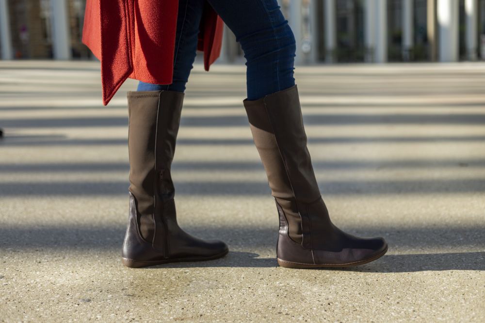 Bekleidet mit kniehohen Stiefeln QATCH Nappa Brown von ZAQQ und Blue Jeans schlendert eine Person über eine sonnenbeschienene Betonoberfläche. Die Schattenmuster erzeugen einen Kontrast auf dem Boden, während sie einen schicken roten Mantel zur Schau stellt.