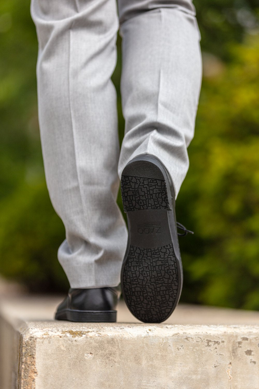 Nahaufnahme einer Person in grauer Hose, die eine Treppe hinaufsteigt. Der Fokus liegt auf ihren schwarzen APEQ-Schuhen von ZAQQ mit einer strukturierten schwarzen Sohle. Der handgefertigte Kalbleder ergänzt die Eleganz. Im Hintergrund ist verschwommenes grünes Laub zu sehen.