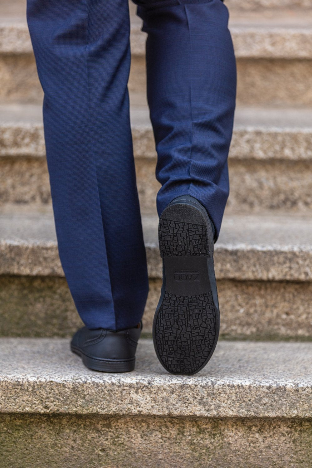 Eine Person in marineblauer Hose und ZAQQs BRIQ Brogue Vegan Black Schuhen geht Steintreppen hinauf. Der Fokus liegt auf der Sohle, die mit einer rutschhemmenden Sohle ausgestattet ist, die beim Aufsteigen für Halt sorgt.