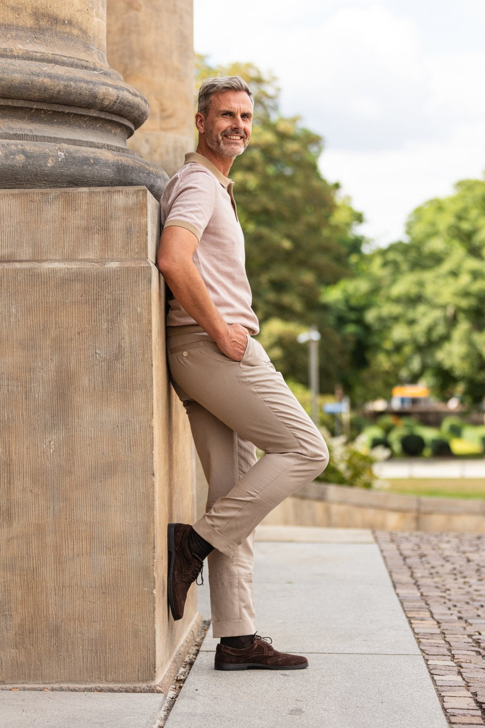 Ein Mann in hellbraunem Hemd und beiger Hose, der stylische ZAQQ BRIQ Brogue Velours Brown-Schuhe trägt, lehnt draußen an einer großen Steinsäule. Er lächelt leicht und sieht entspannt aus, im Hintergrund sieht man Grün und einen bewölkten Himmel.