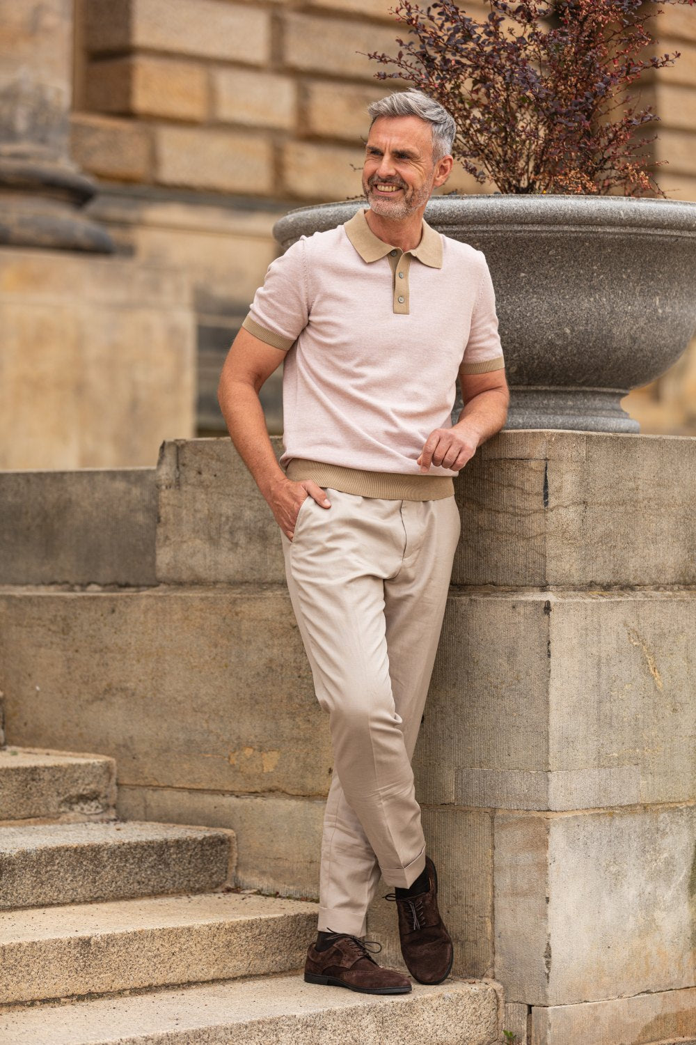 Ein Mann mit grauem Haar steht lässig auf Steinstufen und lehnt an einem großen Steinpflanzgefäß. Er trägt ein hellrosa Poloshirt, beige Hosen und BRIQ Brogue Velours Brown-Schuhe von ZAQQ aus französischem Kalbleder und lächelt freundlich in einer städtischen Umgebung im Freien.