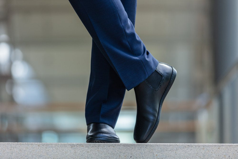 Eine Person in blauen Hosen und schwarzen EQUITY Brogue-Schuhen von ZAQQ steht mit gekreuzten Beinen auf einem Steinvorsprung und wirkt so lässig und doch professionell. Der Hintergrund ist sanft verschwommen, wodurch der Fokus auf die hochwertigen Schuhe und die Kleidung aus Nappaleder gelegt wird.