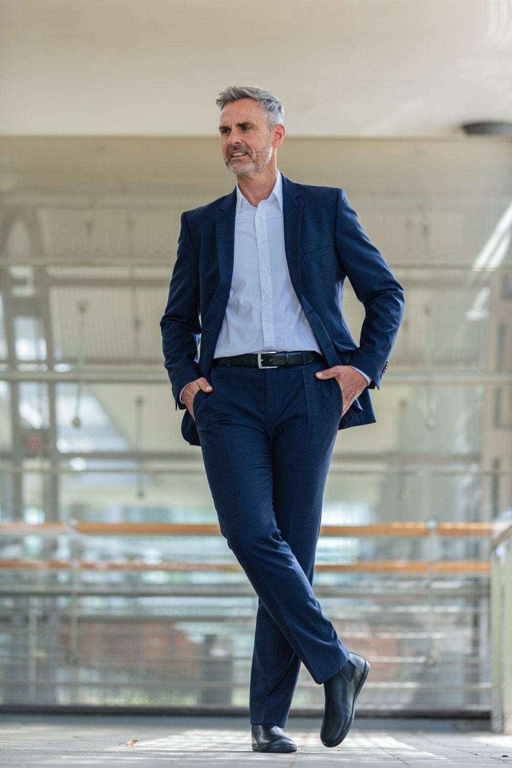 Ein Mann in einem schicken blauen Anzug und weißem Hemd steht selbstbewusst mit den Händen in den Taschen da und schlägt ein Bein übereinander, um seine schwarzen ZAQQ EQUITY Brogue-Schuhe zu präsentieren. Er befindet sich in einem modernen, offenen Raum mit Glas- und Metallarchitektur.