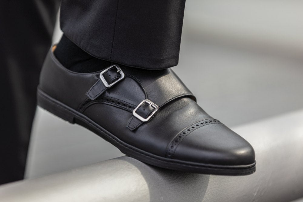 Eine Nahaufnahme des MONQ Captoe Black von ZAQQ strahlt Eleganz aus und zeigt einen schwarzen Monkstrap-Businessschuh aus Leder mit zwei silbernen Schnallen. Der Schuh verfügt über dekorative Perforationen an der Zehenkappe und wird stilvoll mit schwarzen Hosen kombiniert, wobei der Fuß selbstbewusst auf einer Metalloberfläche ruht.