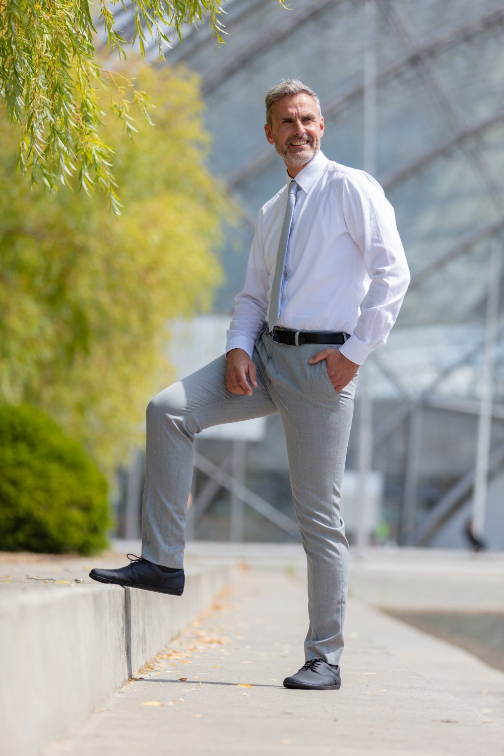 Ein Mann mit kurzen grauen Haaren und Bart lächelt im Freien. Er trägt ein weißes Hemd, eine graue Krawatte und eine graue Hose. Ein Fuß, in einem PEAQ Vegan Black Halbschuh von ZAQQ mit atmungsaktivem Microfaserfutter (atmungsaktives Microfaserfutter), ruht auf einer niedrigen Mauer. Grünflächen und ein Gebäude bilden den Hintergrund dieses ruhigen Moments.