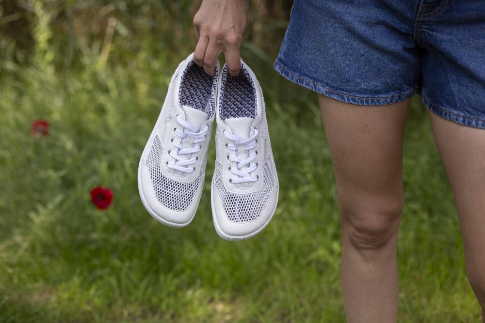 Eine Person in Jeansshorts hält ein Paar weiße ALIQANTE-Sneaker von ZAQQ an den Schnürsenkeln und steht draußen auf grünem Gras mit roten Blumen im Hintergrund. Die Sneaker bieten außergewöhnliche Atmungsaktivität für ultimativen Komfort beim Erkunden der Natur.