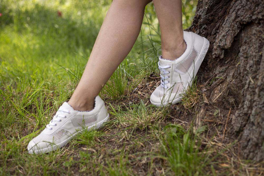 Die Beine einer Person, die weiße ALIQANTE-Sneaker von ZAQQ trägt, lehnen an einem Baumstamm. Diese hochwertigen Sneaker aus Nappaleder bieten hervorragende Atmungsaktivität, da sie auf Gras stehen, umgeben von Grün.