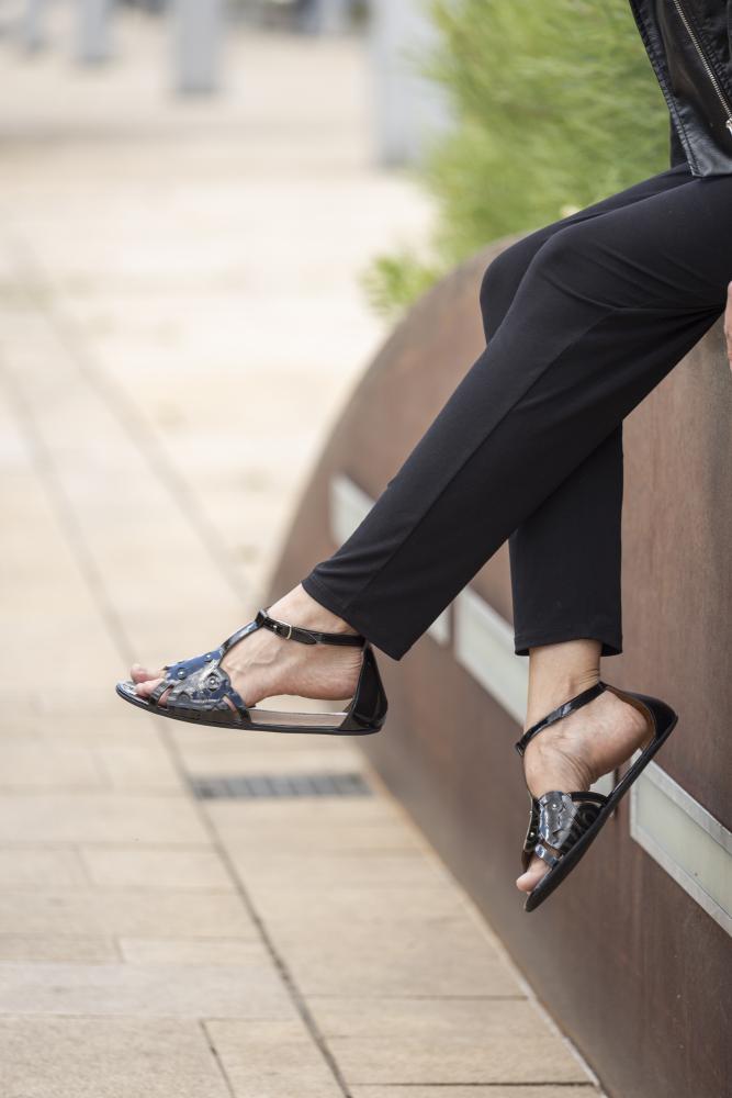 Eine Person sitzt auf einem Felsvorsprung. Sie trägt schwarze Hosen und stylische BOUQET Black Sandalen von ZAQQ, gefertigt aus glattem Lackleder. Im Hintergrund sind ein gepflasterter Gehweg und etwas Grün zu sehen.