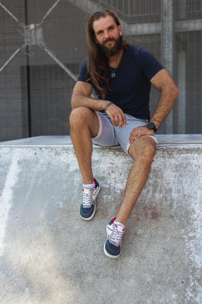 Ein Mann mit langen Haaren und Bart sitzt auf einer Betonkonstruktion. Er trägt ein marineblaues T-Shirt, hellgraue Shorts und dunkelblaue ZAQQ BREAQ-Sneaker. Mit übereinandergeschlagenen Beinen und einer Hand auf dem Knie ruht er auf seinem Gesicht und strahlt einen lässigen und entspannten Ausdruck aus.