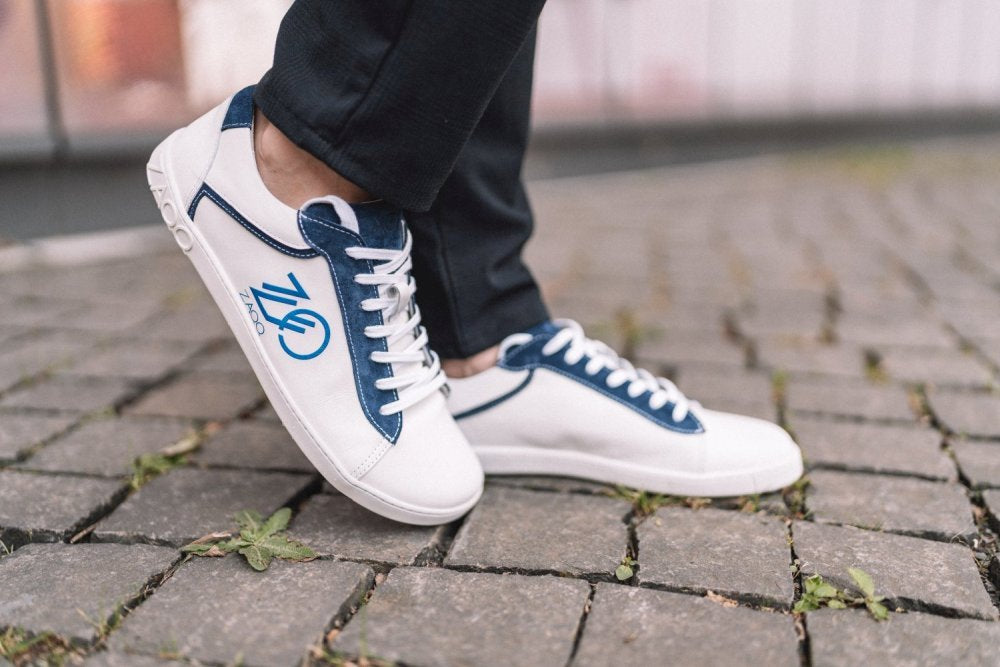 Eine Person, die die blauen CEDRIQ-Sneakers von ZAQQ mit einem runden Logo trägt, schlendert einen Kopfsteinpflasterweg entlang, zwischen den Steinen das Gras hervorschaut.