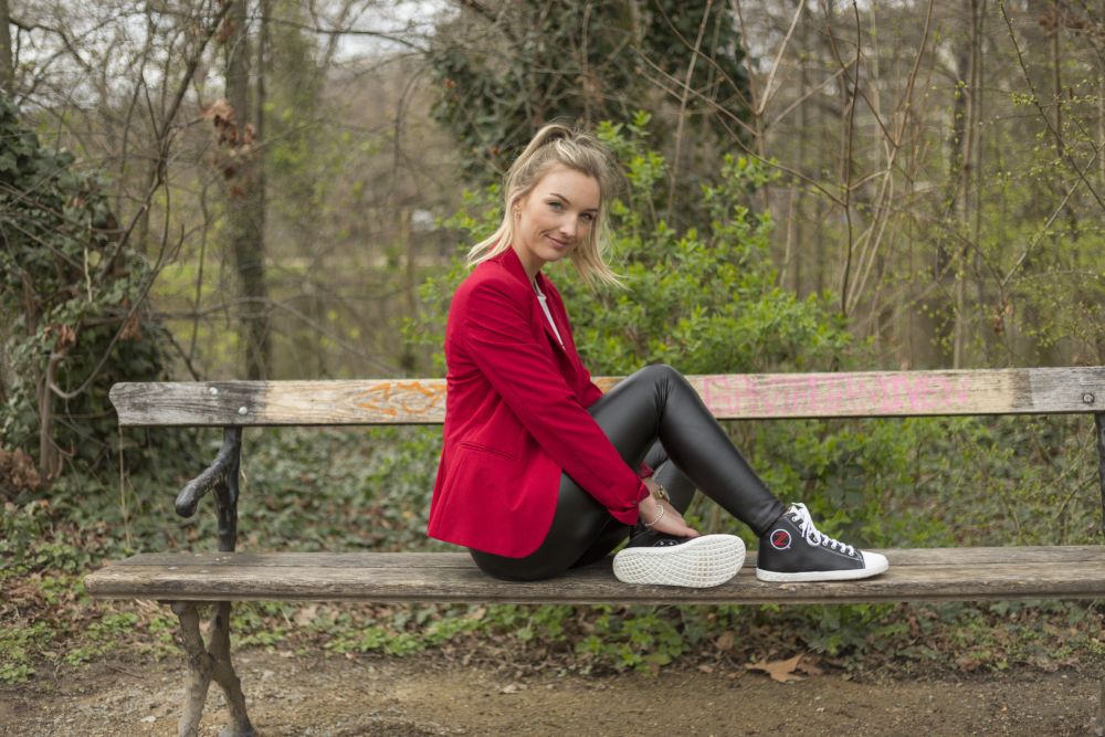 Eine Frau in roter Jacke und schwarzer Lederhose sitzt auf einer Holzbank im Park, lächelt in die Kamera und hält dabei ihre stylischen schwarzen ZAQQ CHUQQS Sneakers, bekannt für ihre bequeme Kautschuksohle. Im Hintergrund sind üppige Bäume und Grünflächen zu sehen.