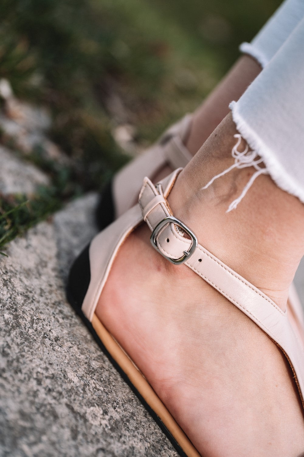 Nahaufnahme einer Person, die CLIQ Nude Riemchensandale-Sandalen von ZAQQ mit Metallschnalle trägt und auf einer Steinoberfläche steht. Die stylischen Sandalen werden mit ausgefransten weißen Jeans kombiniert, was dem Ensemble eine entspannte Ausstrahlung verleiht.