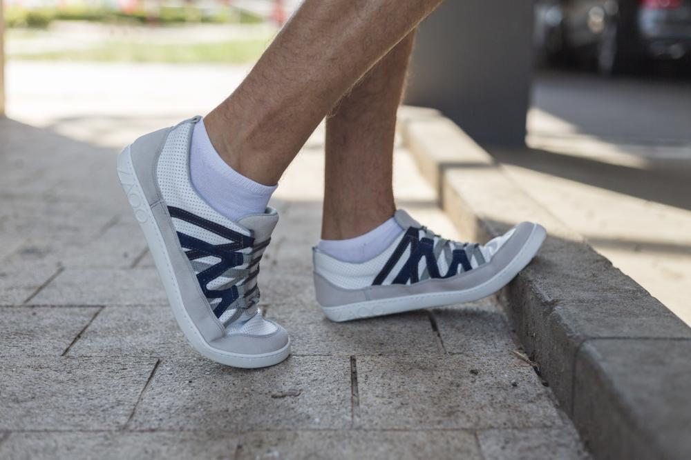 Eine Person, die elegante DRAQE White Grey-Sneakers von ZAQQ mit weißen Socken trägt, steigt von einem Bordstein auf einen gepflasterten Gehweg. Der Hintergrund ist leicht verschwommen und zeigt Bäume und geparkte Autos im Tageslicht, was die hochwertige Nappalederqualität dieser stilvollen Schuhe subtil hervorhebt.