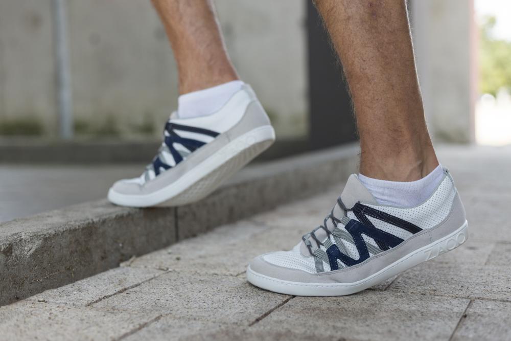 Nahaufnahme einer Person, die weißgraue ZAQQ DRAQE-Sneaker mit blauen Akzenten und weißen Socken trägt und von einem Bordstein steigt. Die Beine und Füße sind teilweise sichtbar, im Hintergrund sind ein Betonweg und ein verschwommenes Gebäude zu sehen.