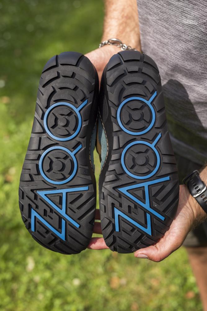 Eine Person steht draußen auf Gras und hält ein Paar wasserfeste Schuhe ESQAPE Blue Orange mit kräftigen schwarzen Profilen und blauen Designs neben dem Wort ZERO. Diese Barfußschuhe von ZAQQ mit ihrem einzigartigen Muster zeigen den TERRA Sohlen-Stil und vereinen Funktionalität und Flair, ideal für jedes Abenteuer.