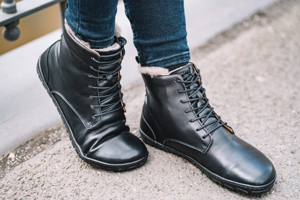 Eine Person in den Schnürstiefeletten QUINTIC Winter Black von ZAQQ mit Lammfell gefüttertem Innenfutter steht auf einem strukturierten Gehweg. Die Stiefel, die sich perfekt als Winterstiefel eignen, werden mit dunklen Jeans kombiniert.