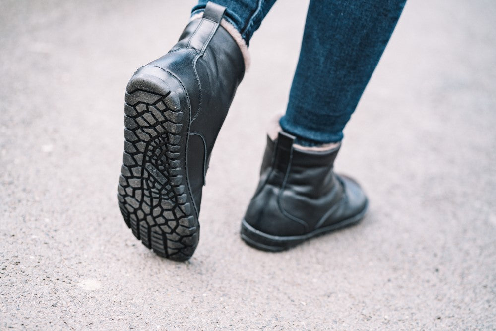 Eine Person läuft auf einem Betonweg und trägt dunkle Jeans und die QUINTIC Winter Black-Stiefel von ZAQQ. Der Fokus liegt auf der Rückseite der Stiefel, wodurch ihr strukturiertes Profilmuster hervorgehoben wird und darauf schließen lässt, dass sie für zusätzliche Wärme mit Lammfell gefüttert sein könnten.