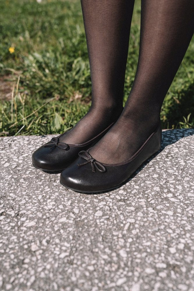 Nahaufnahme der Füße einer Person, die dunkelbraune ZAQQ LOOQ Ballerinas mit Schleifen und schwarze transparente Strumpfhosen trägt. Sie steht auf einem gesprenkelten Bürgersteig, im Hintergrund ist grünes Gras zu sehen.