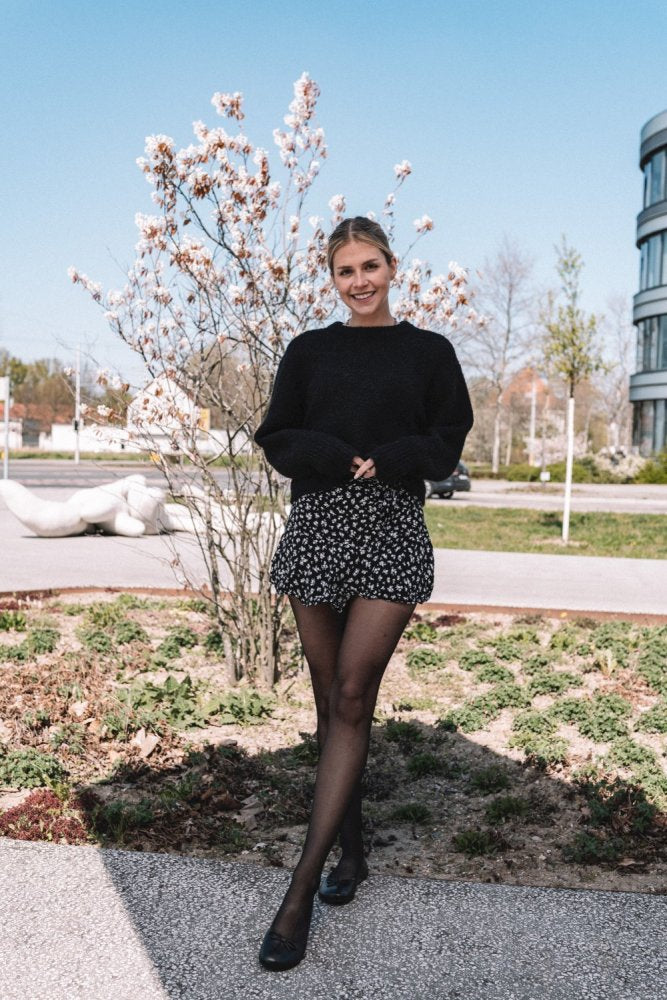 Eine Person steht vor einem blühenden Baum, gekleidet in einen dunkelbraunen LOOQ-Pullover von ZAQQ, dazu einen geblümten Rock und Strumpfhosen. Sie verkörpert die Eleganz einer modernen Barfuß-Ballerina vor der Kulisse eines zeitgenössischen Gebäudes unter einem klaren blauen Himmel.