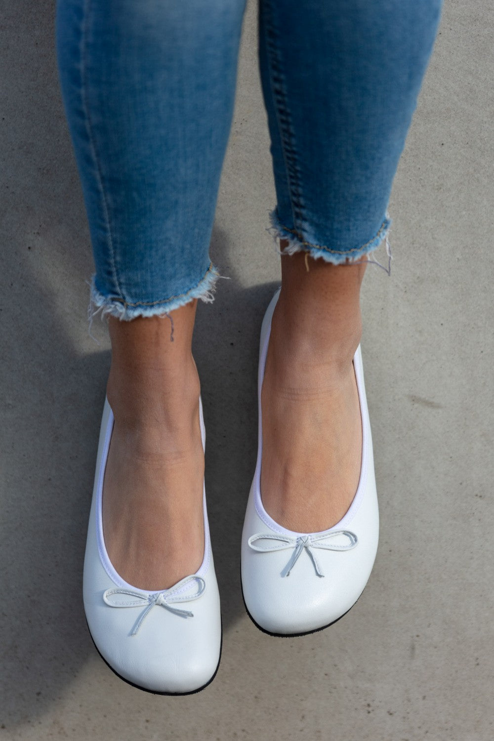 Nahaufnahme einer Person, die Bluejeans mit ausgefranstem Saum und weiße LOOQ-Ballerinas aus Nappaleder von ZAQQ trägt und auf einer Betonfläche steht. Die Ballerinas zeigen ein schlichtes, klassisches Design mit kleinen Schleifen.