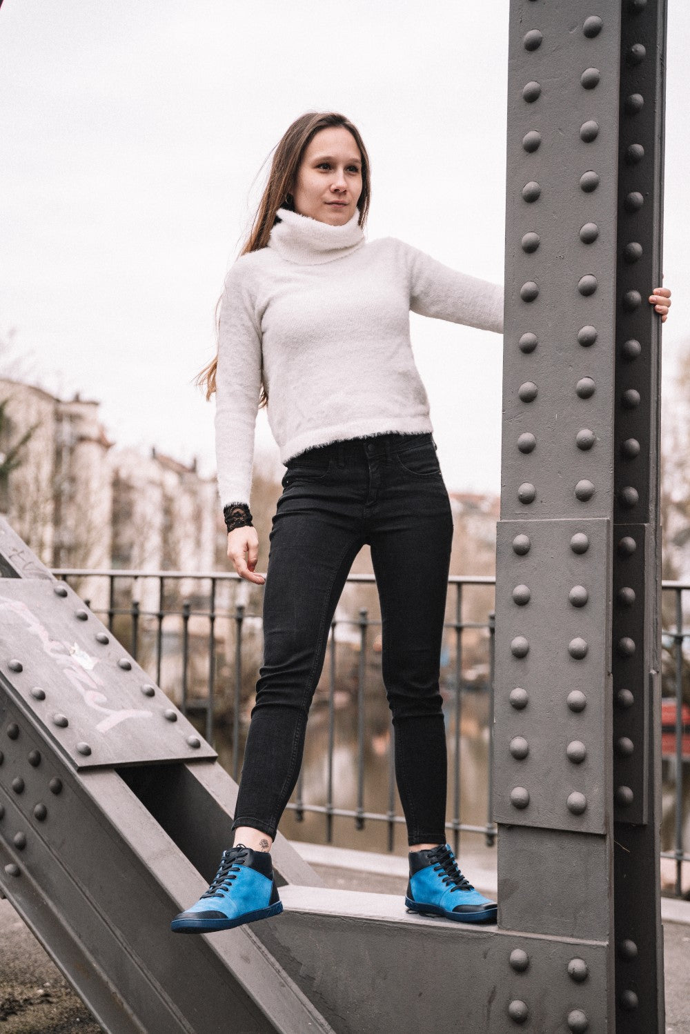 Eine Frau in einem weißen Rollkragenpullover und schwarzen Jeans kombiniert ihr Outfit mit LUQQ Azur-Schuhen von ZAQQ und steht im Freien auf einem Metallträger. Sie hält sich an einem Teil der Struktur fest, im Hintergrund sind Bäume und Gebäude sanft verwischt.