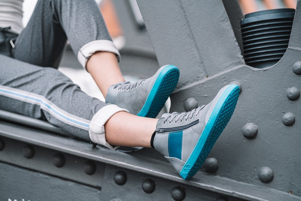 Eine Person, gekleidet in graue Jogginghosen mit hochgekrempelten Hosenbeinen und stylische ZAQQ LUQQ Turquoise Sneakers mit blauer Sohle und Akzenten, sitzt auf einer Metallkonstruktion, die an eine Brücke oder ein Industriegelände erinnert und eine entspannte, urbane Atmosphäre vermittelt.