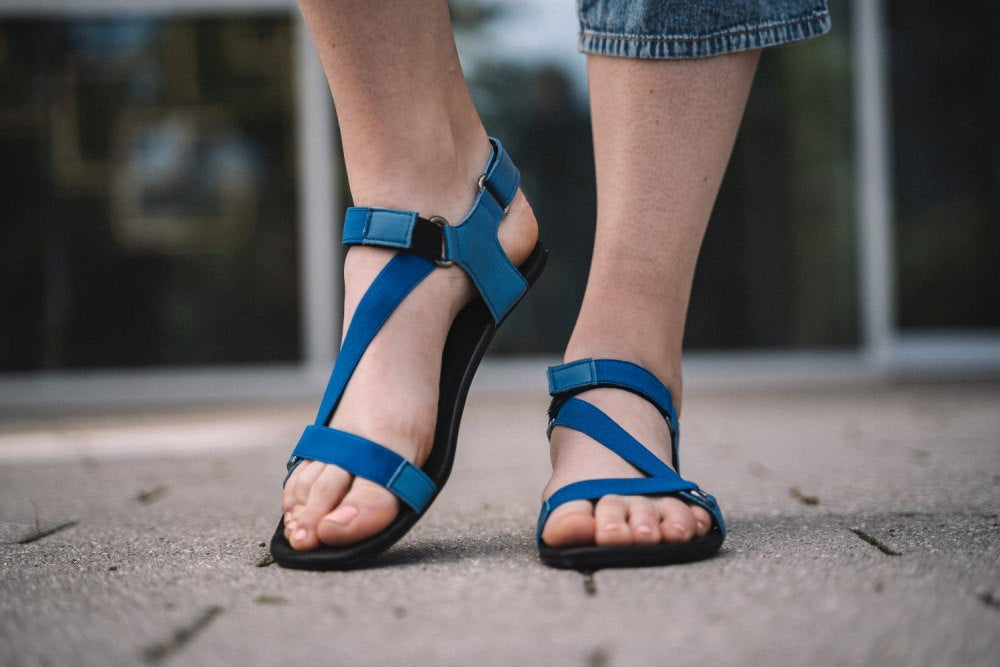 Eine Person, die die MIQA Blue Barfußsandalen von ZAQQ trägt, steht auf einer Betonfläche. Ihre blauen Riemchenschuhe sind mit Klettverschlüssen sicher befestigt. Sie trägt verkürzte Jeanshosen, während der Hintergrund kunstvoll verschwommen ist, um die stylischen Sandalen hervorzuheben.