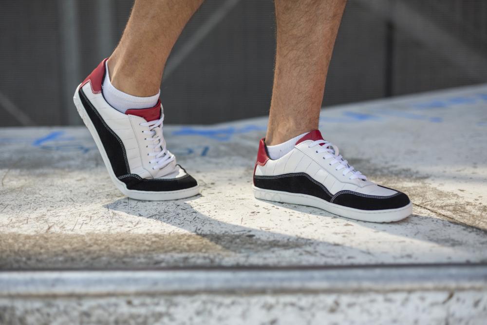 Nahaufnahme einer Person, die in den QAMPION Black White-Sneakers von ZAQQ mit schwarzen und roten Akzenten und einer flexiblen Sohle schreitet. Der Fokus liegt weiterhin auf dem Schuhwerk, das aus glattem Veloursleder gefertigt ist, wobei nur die Beine unterhalb der Knie vor der kahlen Betonoberfläche sichtbar sind.