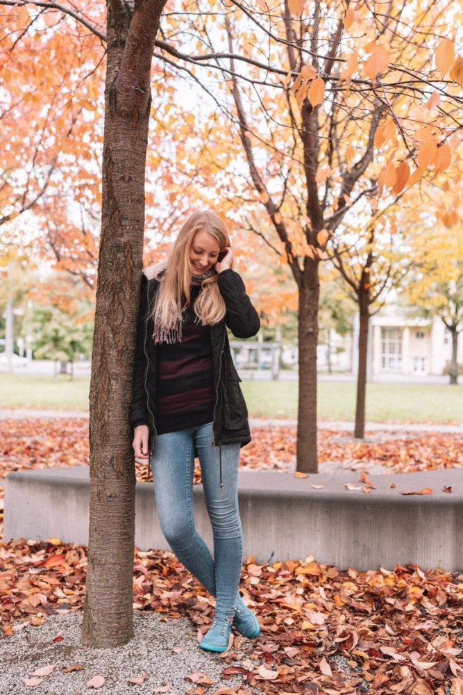 Eine Frau mit langen blonden Haaren steht in einem Park an einem Baum, umgeben von Herbstlaub. Sie trägt eine dunkle Jacke, Jeans und die Schuhe QARLY Turquoise von ZAQQ. Die Bäume um sie herum haben orange-gelbes Laub und der Boden ist mit Laub bedeckt.