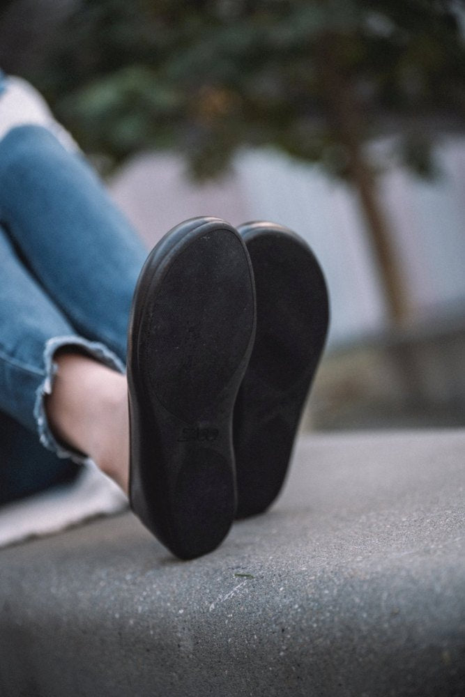 Eine Nahaufnahme einer Person in Blue Jeans, die auf einer Betonfläche sitzt und die QARMA-Sohlen ihrer dunkelbraunen ZAQQ-Schuhe zeigt. Der Hintergrund ist sanft verschwommen und weist einen Hauch von Grün auf.