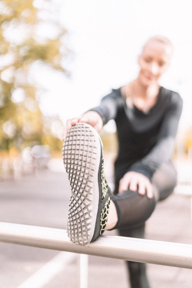 Eine Frau in Sportkleidung streckt ihr Bein auf einem Metallgeländer im Freien aus und präsentiert die Sohle ihres QASEY Green-Schuhs von ZAQQ. Der Schuh besteht aus atmungsaktivem Netzgewebe und veganen Materialien und strahlt vor einem verschwommenen Hintergrund aus Bäumen im hellen Tageslicht Komfort und Umweltfreundlichkeit aus.