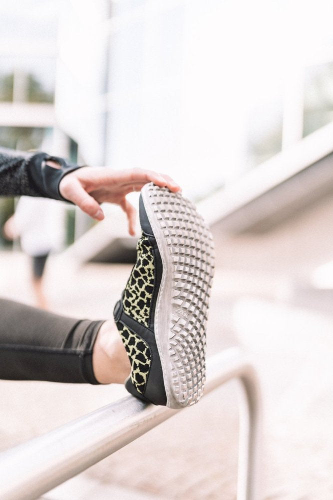 Eine Person streckt ihr Bein auf einem Metallgeländer aus und präsentiert den QASEY Green Sneaker von ZAQQ mit einem schwarz-gelben Animalprint-Design aus veganen Materialien. Die strukturierte Sohle ergänzt das atmungsaktive Mesh-Gewebe und der sanft verschwommene Hintergrund lässt auf eine urbane Umgebung schließen.