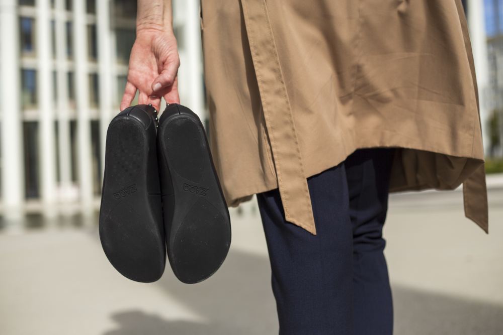 Eine Person in einem braunen Mantel trägt ein Paar ZAQQ DIEQO Nappa Black-Schuhe an den Absätzen und geht an einem sonnigen Tag im Freien spazieren. Die natürliche Fußbewegung wird gefördert, während sie an einem hellen Gebäude mit hohen Säulen vorbeischlendert.