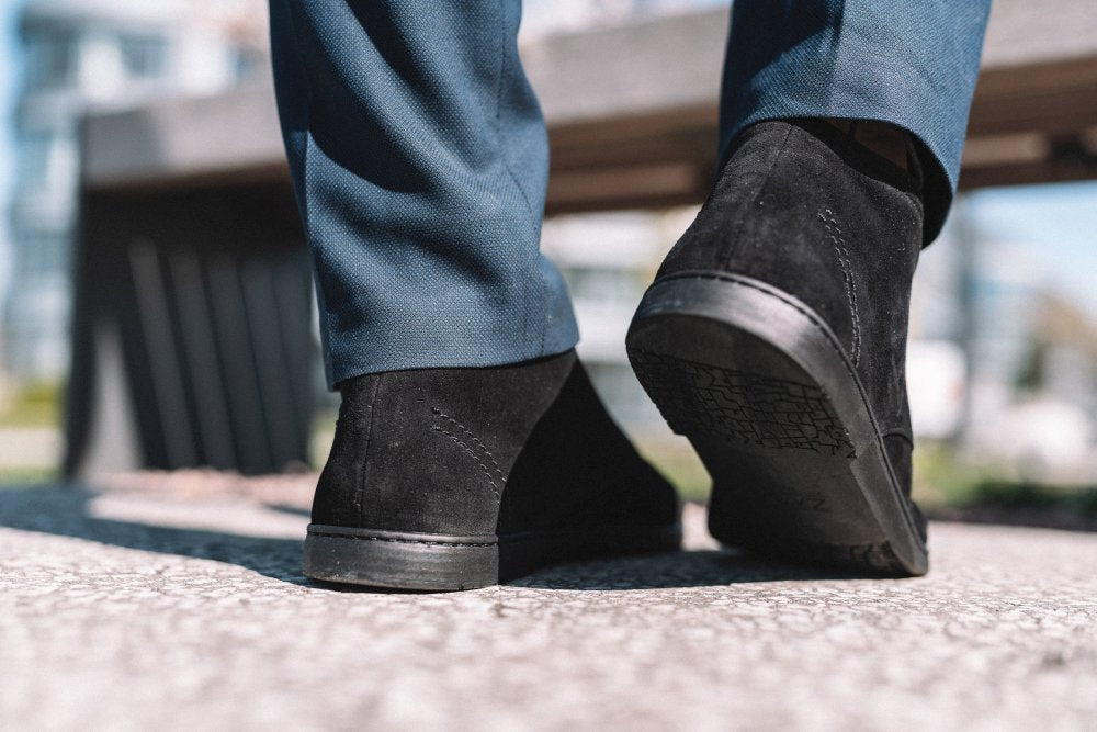 Eine Nahaufnahme zeigt eine Person, die stylische schwarze QLASSY-Velourslederschuhe von ZAQQ und blaue Hosen trägt und selbstbewusst über einen Betonweg schreitet. Der verschwommene urbane Hintergrund lässt das Stadtleben erahnen, komplett mit einer Bank an der Seite.