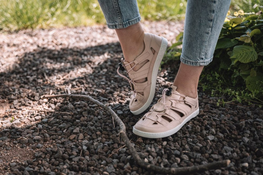 Eine Person, die ZAQQs QLEAR Light Pink-Sneaker mit Cut-Out-Design und Blue Jeans trägt, steht auf einem Kiesweg. Auf dem Boden liegt ein Holzstock für natürlichen Halt, im Hintergrund sind grüne Blätter zu sehen.