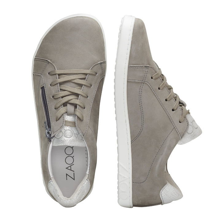 Wir präsentieren die QOMFY Wide Grey Sneaker von ZAQQ: ein Paar graue Sneaker aus Veloursleder mit seitlichen Reißverschlüssen und weißen Sohlen. Ein Schuh ist von oben zu sehen, wodurch die Schnürsenkel und das Logo auf der Innenseite hervorgehoben werden, während der andere Schuh aufwendige Nähte und Designelemente für einen stilvollen und dennoch bequemen Look aufweist.