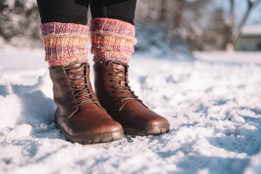 Eine Nahaufnahme von jemandem, der QUINTIC Winter Vegan Nut-Lederstiefel von ZAQQ in Braun trägt, kombiniert mit farbenfrohen Strick-Stulpen. Sie stehen im Schnee, mit einer verschneiten Landschaft und einem blauen Himmel, die im Hintergrund sanft verschwommen sind.