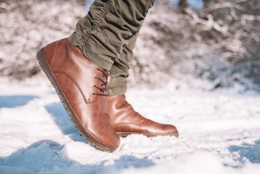 Eine Person in QUINTIC Winter Vegan Nut-Stiefeln von ZAQQ schreitet durch den Schnee. Sie trägt grüne Hosen vor einem winterlichen Hintergrund mit verschwommenen Bäumen, wodurch ihr veganes Schuhwerk und der Schnee darunter betont werden.