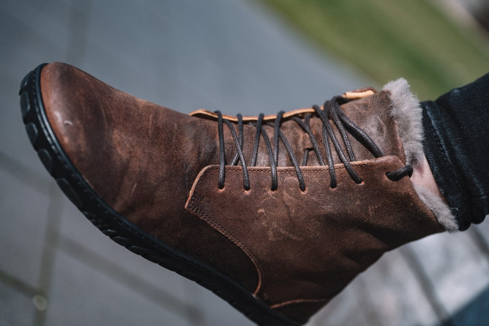 Eine Nahaufnahme zeigt einen Fuß in der QUINTIC Winter Velours Brown Waterproof Lederstiefelette von ZAQQ mit schwarzen Schnürsenkeln und Gummisohle. Das Lammfell-Futter lugt oben hervor und sorgt für behagliche Wärme. Im Hintergrund verschwimmen ein gefliester Gehweg und Gras, während dunkle Hosen den Look vervollständigen.