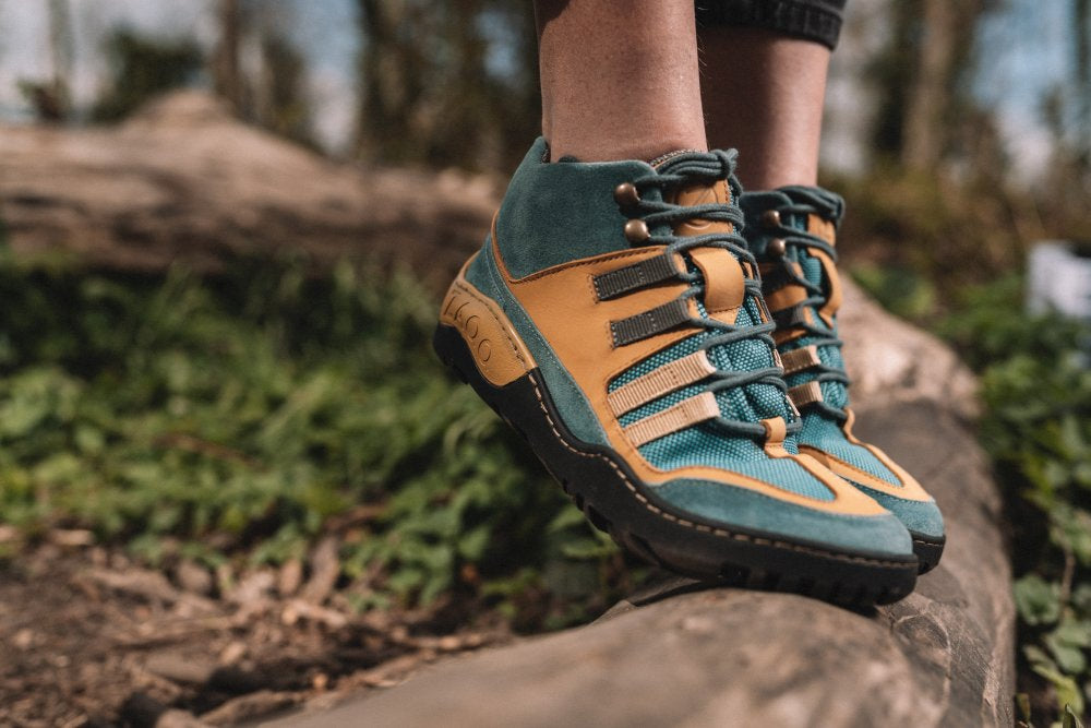 Eine Person, die die SQOUT Blue Orange Waterproof-Schuhe von ZAQQ trägt, steht auf einem Baumstamm. Diese Schuhe im Barfußschuh-Stil haben robuste Sohlen und dicke Schnürsenkel und sind daher perfekt zum Wandern geeignet. Ihre atmungsaktiven (atmungsaktiv) und wasserdichten (wasserdicht) Eigenschaften sorgen für Komfort bei jedem Wetter. Der Hintergrund ist verschwommen und lässt Grünflächen und Bäume erkennen.