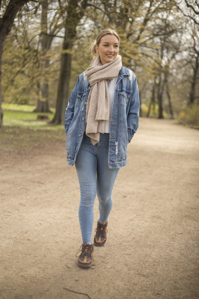 Eine Frau spaziert auf einem Feldweg in einem Park entlang, umgeben von hohen Bäumen. Sie trägt eine Jeansjacke, blaue Jeans, einen beigen Schal und braune SQOUT Brown Waterproof-Stiefel von ZAQQ, lächelt und wirkt entspannt. Die Szene ist ruhig und natürlich.