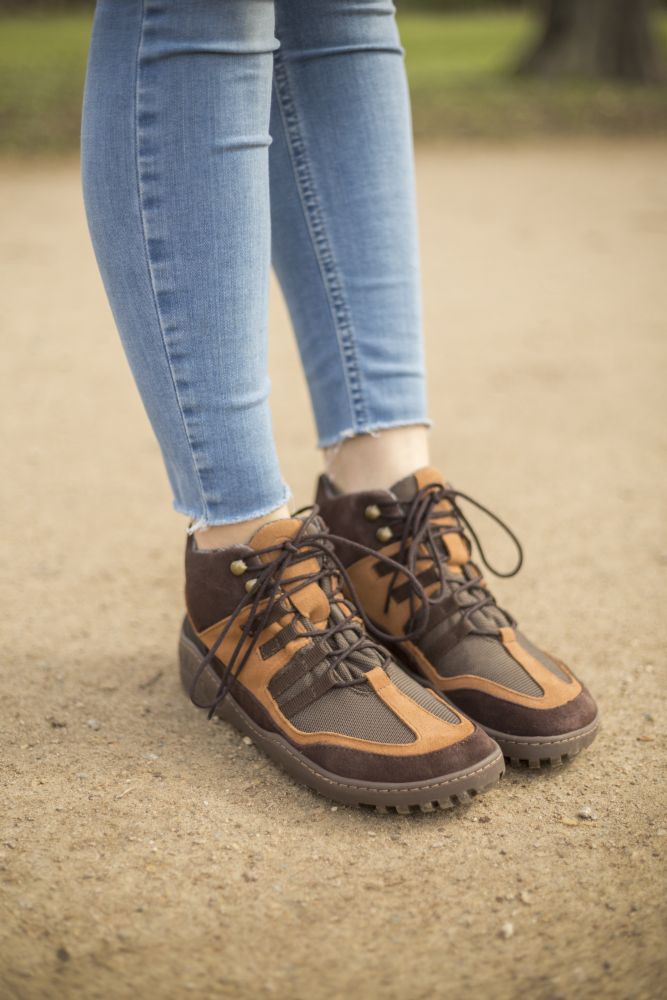 Eine Nahaufnahme einer Person, die hellblaue Jeans und braune Wanderstiefel aus Veloursleder trägt, identifiziert als ZAQQ SQOUT Brown Waterproof. Diese Stiefel verfügen über ein Schnürdesign und eine ZAQQ TERRA-Sohle, die Wildleder und Mesh-Materialien gekonnt kombiniert und so das ideale wasserdichte Schuhwerk für Outdoor-Abenteuer darstellt.