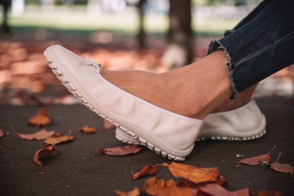 Nahaufnahme der Füße einer Person, die die weißen Mokassinschuhe SUQAR von ZAQQ trägt, die aus geschmeidigem Nappaleder gefertigt sind und eine flexible Sneakersohle haben, während sie auf einem mit Herbstlaub bedeckten Gehweg läuft. Die Person trägt dunkelblaue Jeans mit ausgefransten Säumen.