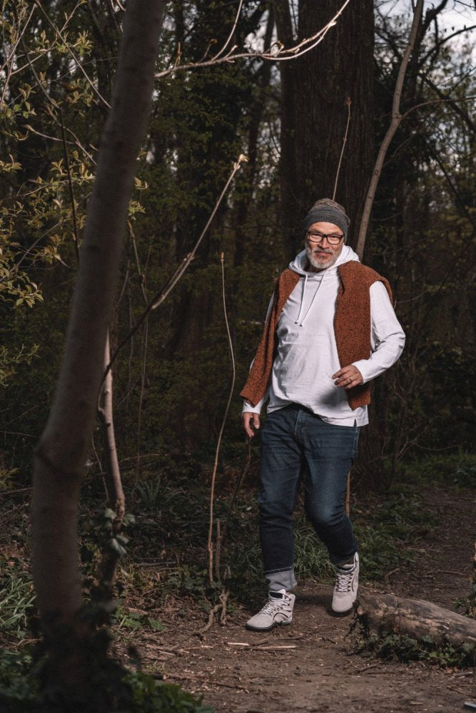 Ein Mann in einer braunen Weste, einem weißen Kapuzenpullover, Jeans, wasserdichten Turnschuhen aus der TERRAQ Beige-Kollektion von ZAQQ und einer Beanie-Mütze schlendert einen Waldweg entlang, der von hoch aufragenden Bäumen und üppigem Grün umgeben ist.