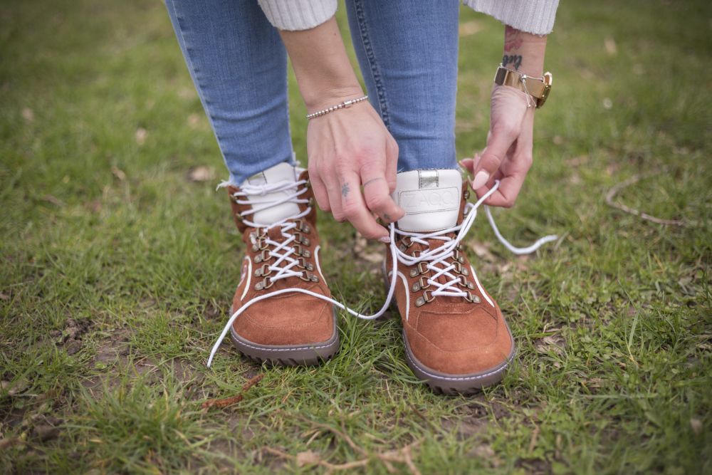 Auf dem grünen Gras kniet eine Person und schnürt die Schnürsenkel ihrer ZAQQ TREQ Cognac Waterproof Wanderschuhe. Sie trägt hellblaue Jeans und einen weißen Pullover und vervollständigt ihren Look mit einem Armband und einer Uhr an ihren Handgelenken.