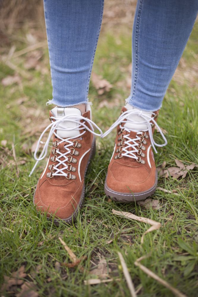 Eine Person in Blue Jeans und den wasserdichten Wanderschuhen TREQ Cognac von ZAQQ steht auf grünem Gras, auf dem trockene Blätter verstreut liegen. Die wasserdichten Stiefel haben weiße Schnürsenkel und Metallösen.