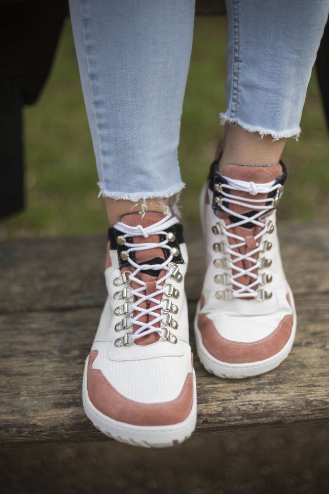 Eine Nahaufnahme zeigt eine Person, die auf einer Holzbank sitzt und die wasserdichten VAQATION Rosa High-Top-Sneakers von ZAQQ trägt, die mit detaillierten Metallösen verziert sind. Ihre hellblauen Jeans haben ausgefranste Säume, was die lässige Atmosphäre des verschwommenen Hintergrunds verstärkt.