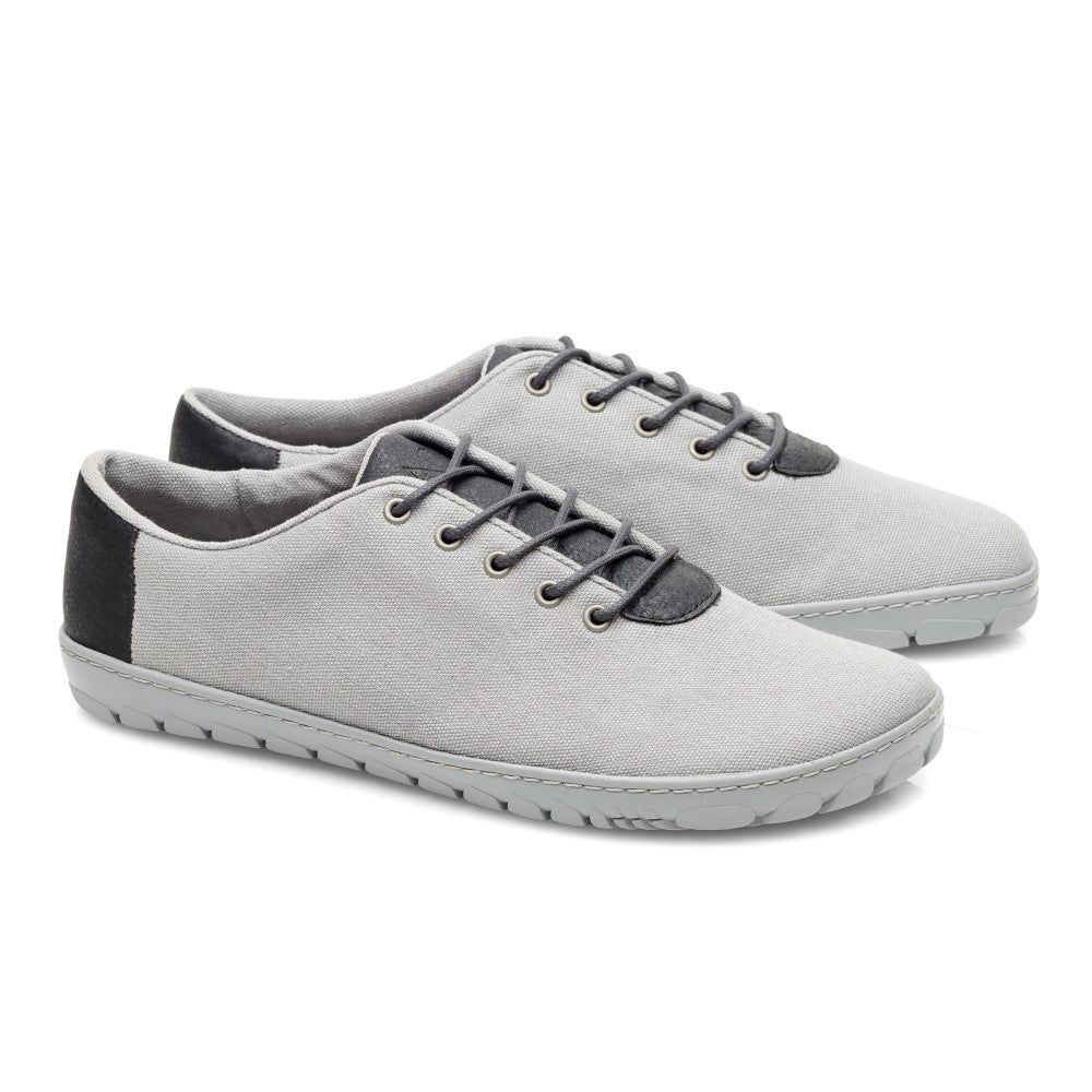 Ein Paar QARO Black Grey Casual-Sneaker von ZAQQ, in Hellgrau mit dunkelgrauen Akzenten und Schnürsenkeln auf weißem Hintergrund. Diese veganen Barfußschuhe zeichnen sich durch ein minimalistisches Design mit Low-Top-Silhouette und Gummisohlen aus und bieten sowohl Stil als auch Komfort.