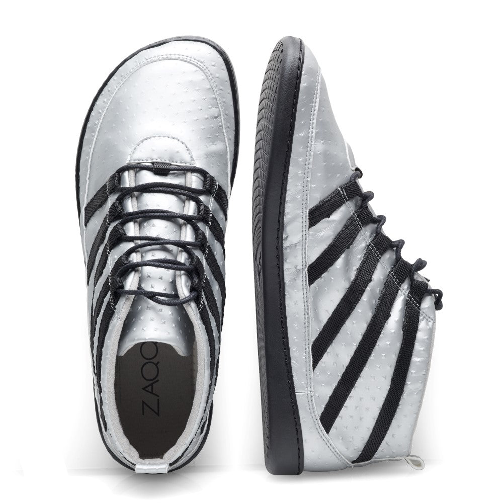 Die SPARQ Mid Silver Sneaker von ZAQQ sind ein Paar silberne High-Tops mit schwarzen Schnürsenkeln und eleganten schwarzen Seitenstreifen auf weißem Hintergrund. Hergestellt aus veganem Obermaterial, ist der linke Sneaker von der Seite zu sehen, während der rechte von oben betrachtet wird und diese schicken Barfußschuhe in Szene setzt.